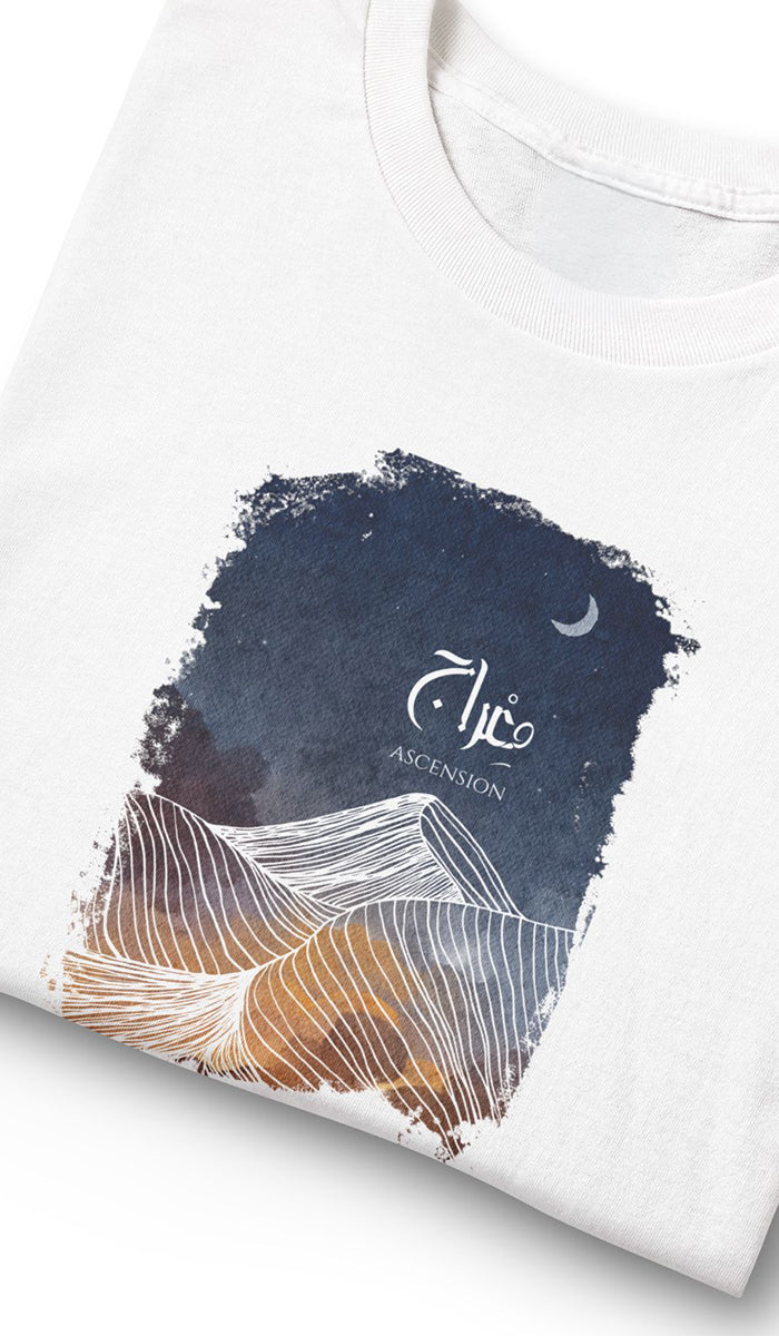 A folded white t-shirt featuring a graphic print with a dark navy blue textured background, white Arabic calligraphy reading 'Ascension', and stylized desert dune illustrations in white and warm brown tones.