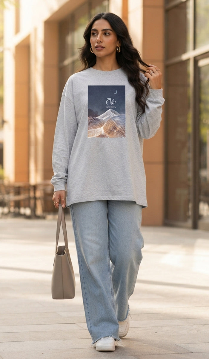 A woman walking outdoors wearing a light gray long-sleeve graphic sweatshirt with a mountain print, light blue wide-leg jeans, white sneakers, and carrying a beige tote bag.