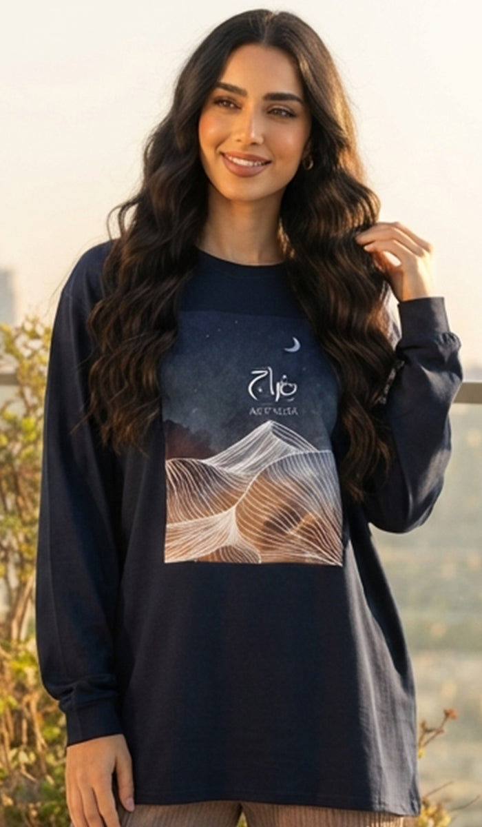 A woman wearing a navy blue long-sleeve tunic top with a graphic print of mountains under a starry night sky with Arabic calligraphy, set against a blurred outdoor background.