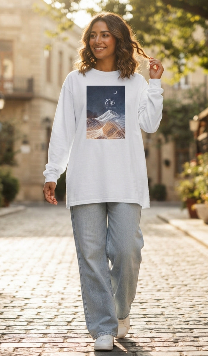 A woman walking on a cobblestone street wearing a white long-sleeve t-shirt with a square graphic print of a mountain and the number 12, paired with light blue jeans.