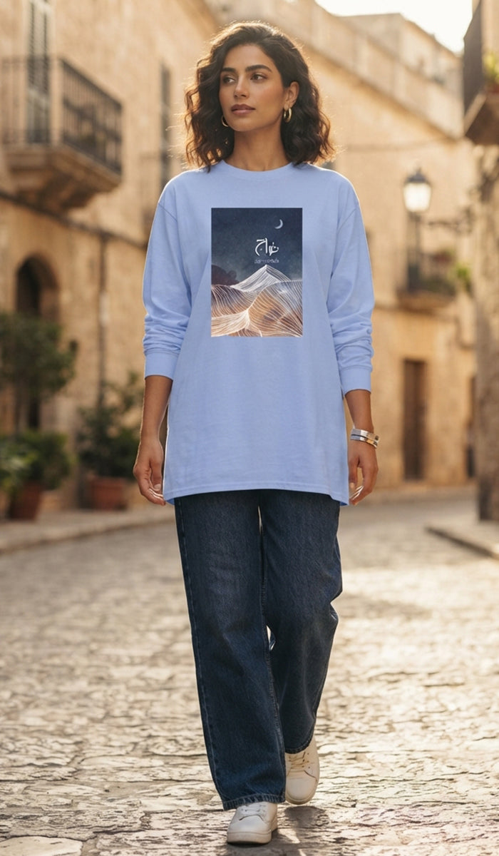 A woman walking down a cobblestone street wearing a light blue long-sleeve t-shirt with a graphic print of a mountain under a starry sky, paired with dark blue jeans and white sneakers.