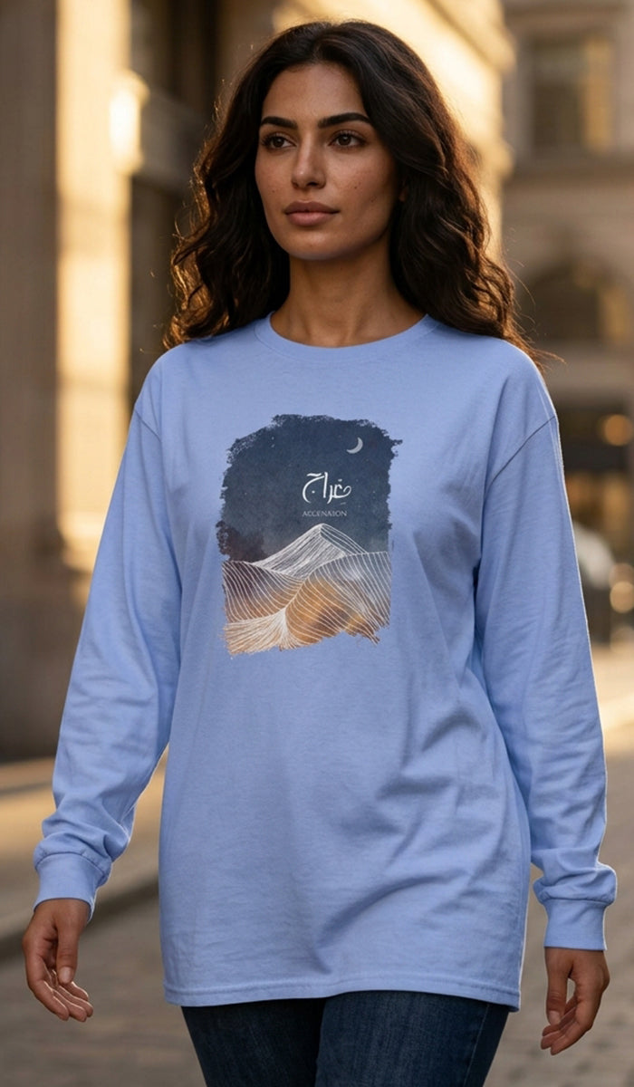 A woman wearing a light blue long-sleeve t-shirt with a graphic print of a mountain under a night sky, standing in an outdoor urban setting.