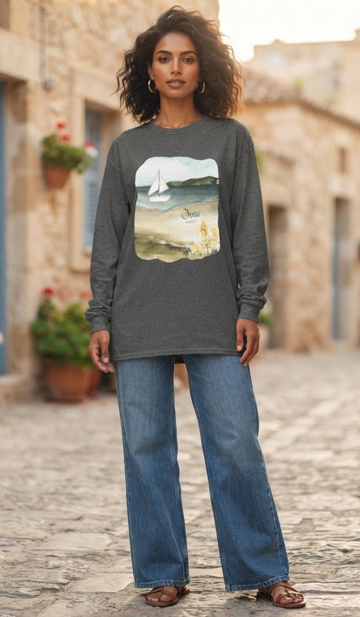 A woman modeling a dark gray long-sleeve t-shirt with a scenic landscape graphic on the front, paired with wide-leg blue jeans and brown sandals, standing on a cobblestone street.