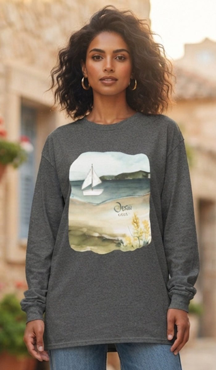 A woman wearing a dark gray long-sleeve t-shirt with a square graphic print of a sailboat on a beach. The background shows a softly blurred outdoor setting with warm stone buildings.