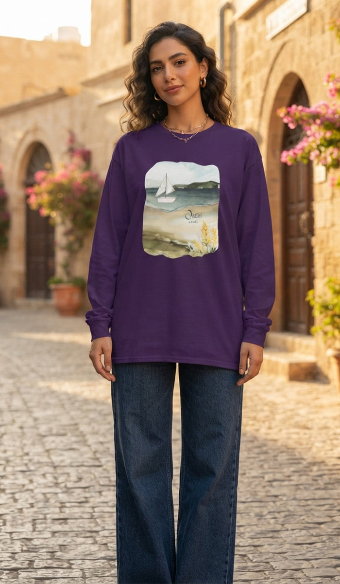 A woman modeling a purple long-sleeve crewneck sweatshirt with a central graphic print of a sailboat on water, paired with dark blue jeans, standing on a cobblestone street in front of a stone building.