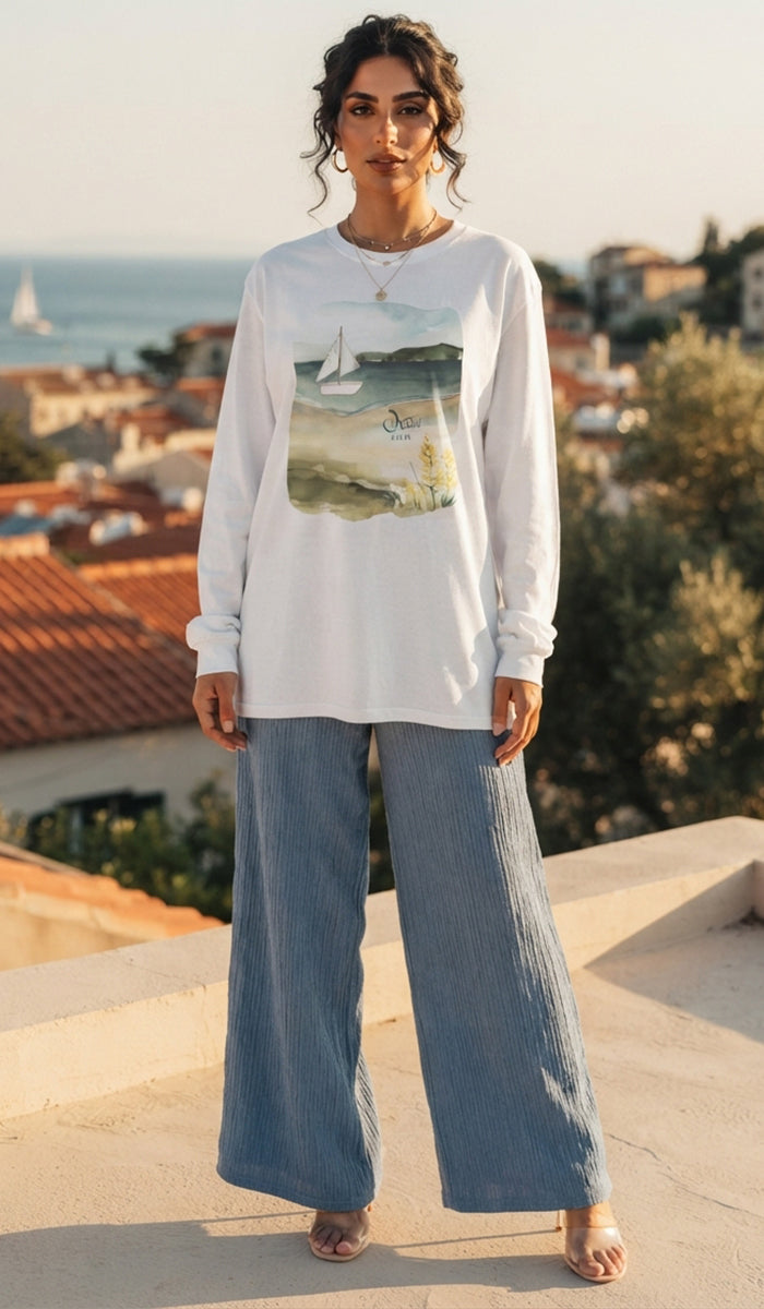 A woman stands outdoors wearing a white long-sleeve graphic T-shirt with a coastal print and wide-leg blue jeans, with a coastal town and sea in the background.