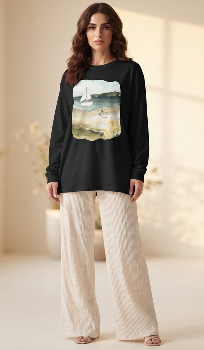 A woman modeling a black long-sleeve t-shirt with a large square graphic print of a sailboat on water, paired with cream-colored wide-leg pants in a brightly lit indoor setting.