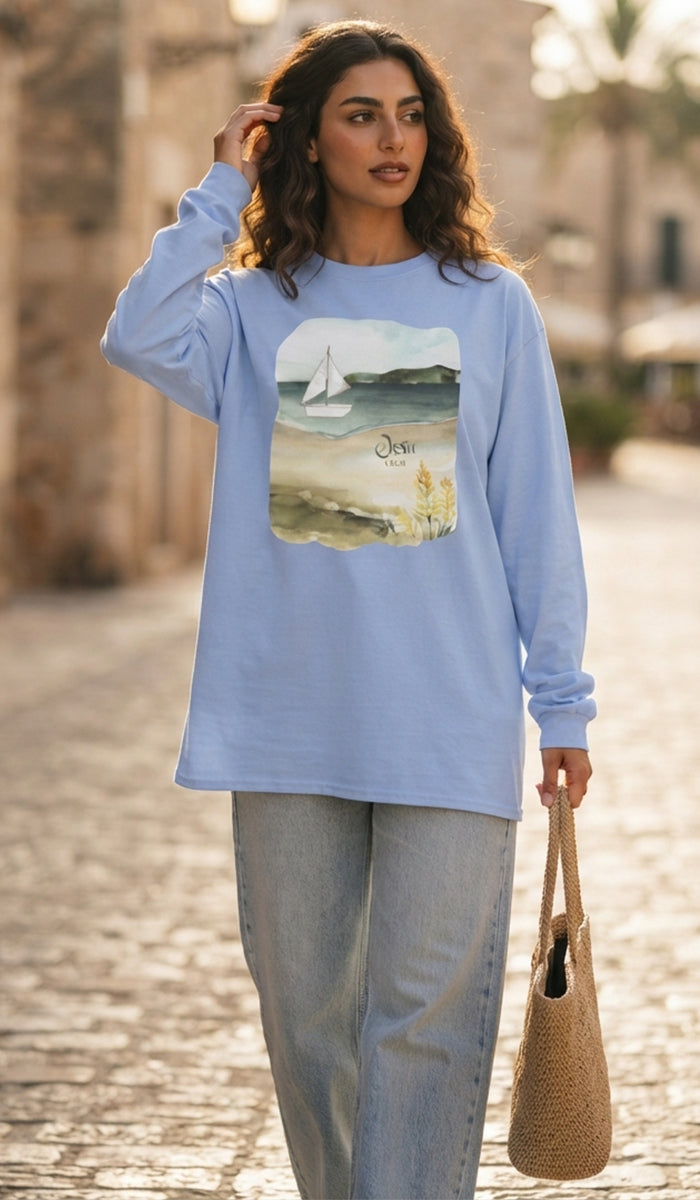 A woman wearing a light blue long-sleeve t-shirt with a square beach graphic print on the front, paired with light-wash jeans and holding a woven straw bag, set against a blurred cobblestone street background.