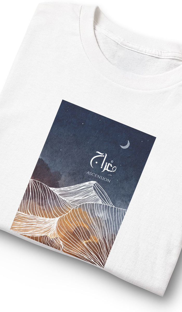 A folded white t-shirt with a rectangular graphic print on the front. The print features a dark navy blue night sky with a crescent moon and white Arabic calligraphy, layered over stylized desert dunes in shades of brown and gold.