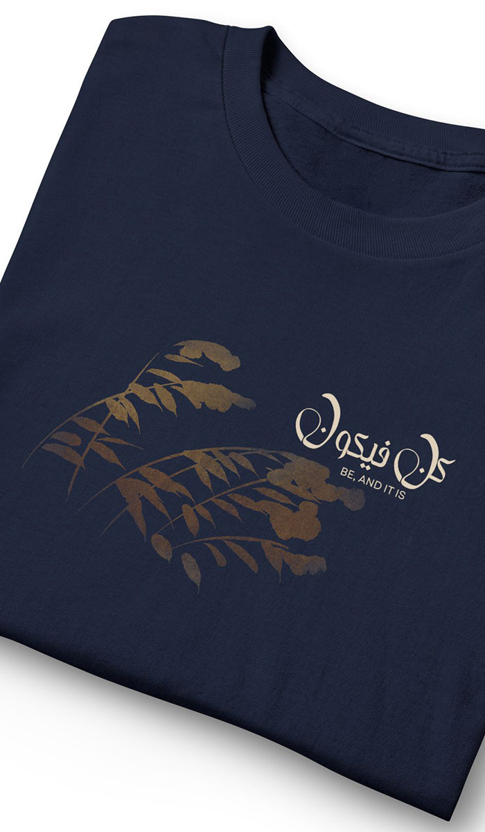A folded navy blue t-shirt with a gold fern leaf graphic and white Arabic calligraphy reading 'Be And It Is' on the front, set against a plain white background.