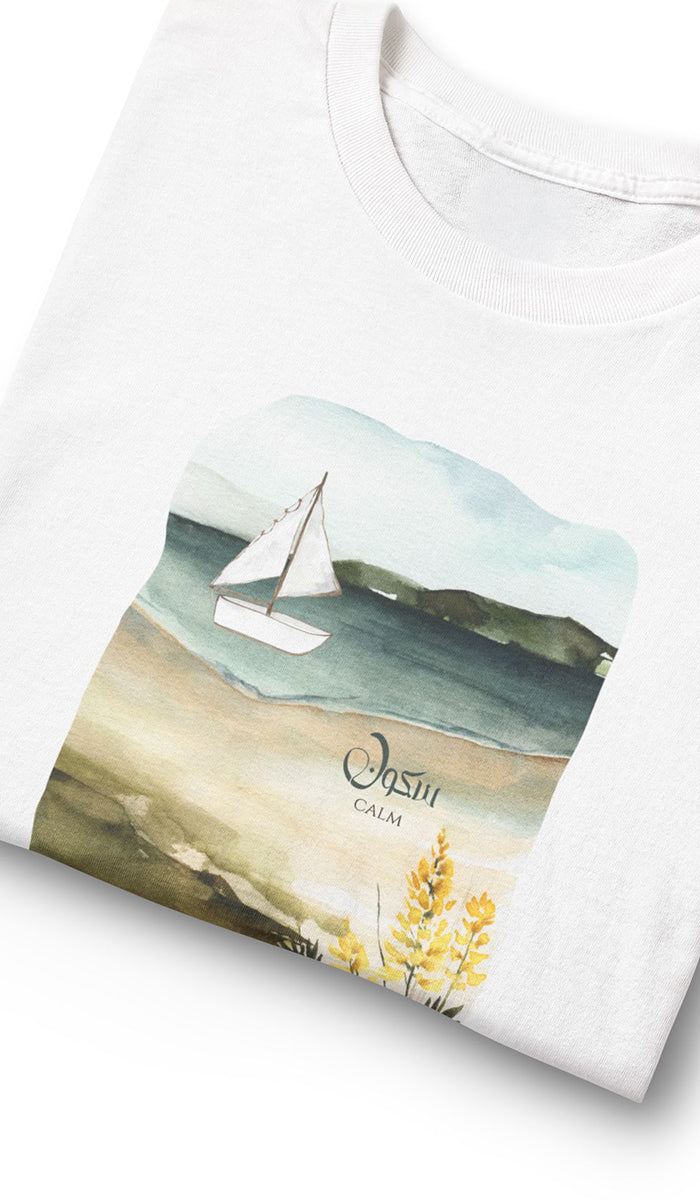 A folded white t-shirt featuring a watercolor-style graphic print of a sailboat on a beach with the text 'Oslo CALM'.