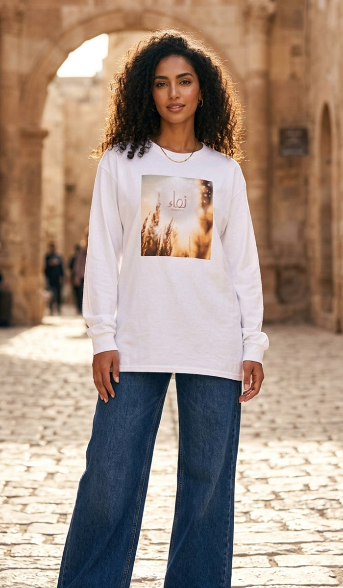 A woman wearing a white long-sleeve t-shirt with a square graphic print on the front, paired with wide-leg blue jeans, standing in front of a stone archway.