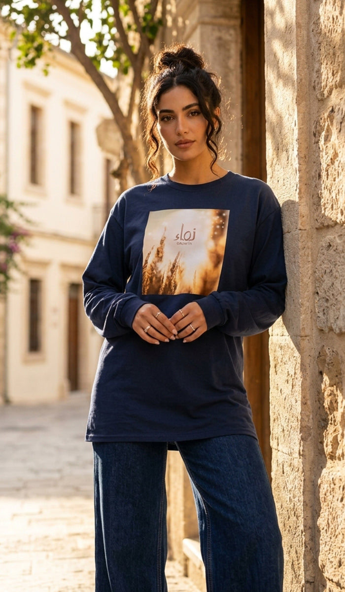 A woman wearing a navy blue long-sleeve t-shirt with a square graphic print on the front, paired with dark blue jeans, standing outdoors against a rustic stone wall.