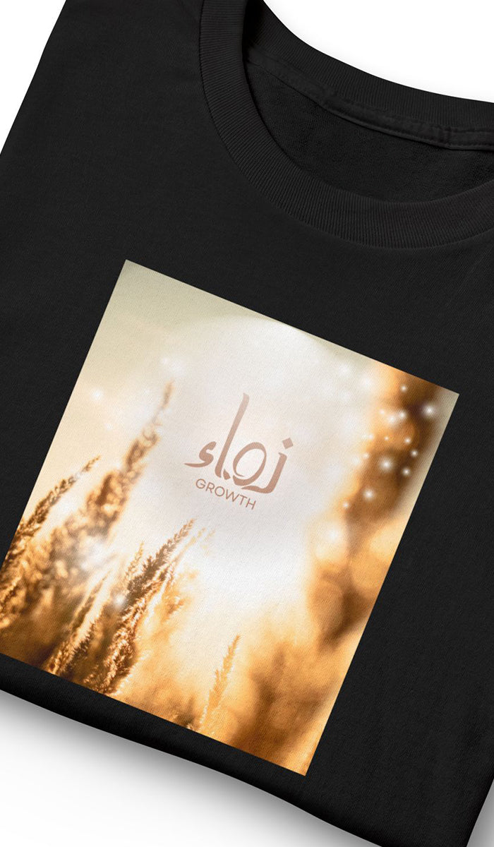 A close-up of a black t-shirt featuring a square graphic print on the front. The print displays a warm, golden wheat field with bokeh light effects and the Arabic word 'Nama' with 'GROWTH' written underneath in a light brown font.
