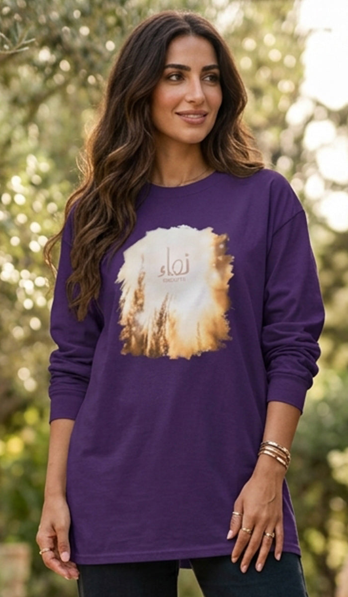 A woman wearing a purple long-sleeve t-shirt with a large graphic print of a desert sunset and Arabic calligraphy, set against a blurred green outdoor background.