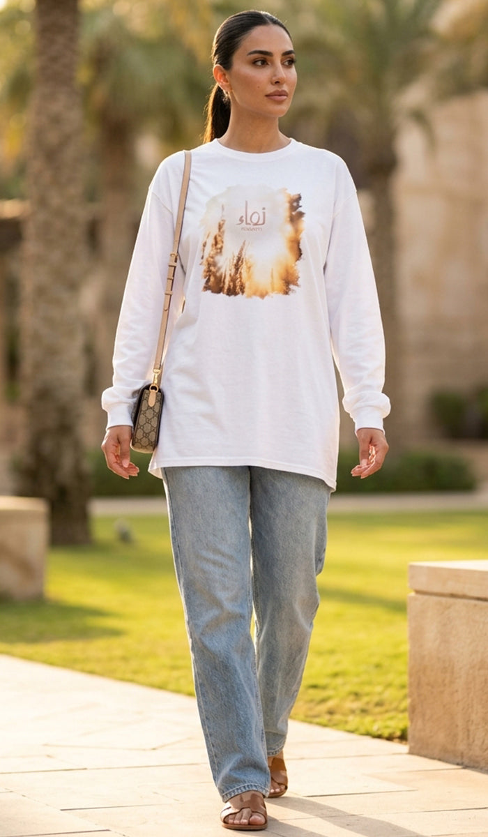 A woman walking outdoors wearing a white long-sleeve graphic T-shirt with a desert sunset print, light blue jeans, and carrying a small crossbody bag.