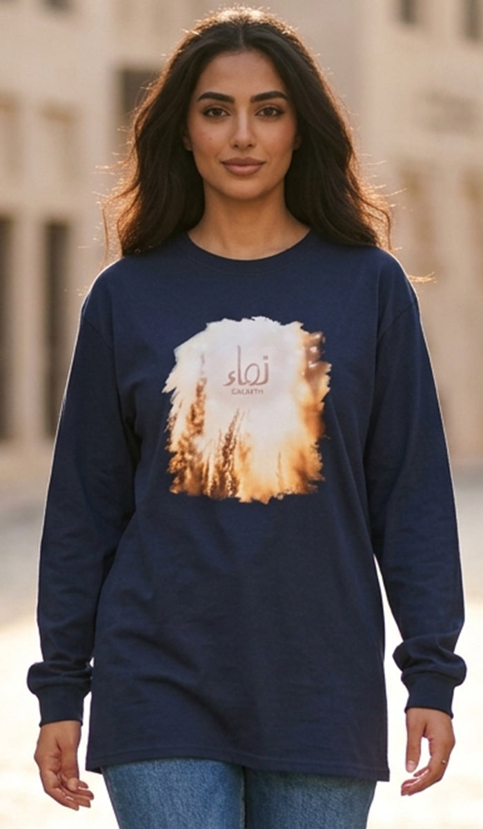 A woman wearing a navy blue long-sleeve t-shirt with a circular graphic print of a sunset over wheat fields and the Arabic word 'Najwa' in the center, paired with blue jeans.