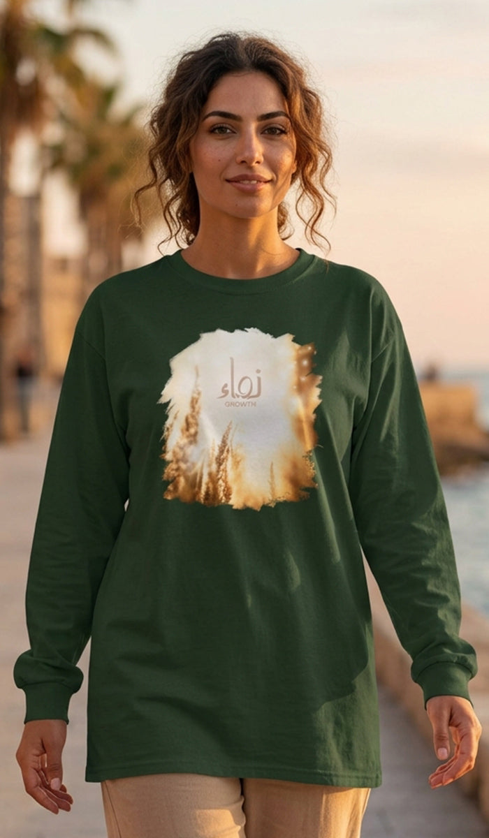 A woman wearing a dark green long-sleeve t-shirt with a circular graphic print of a desert landscape and the Arabic word 'Tawhid' in the center, standing outdoors on a sunny beachside path.