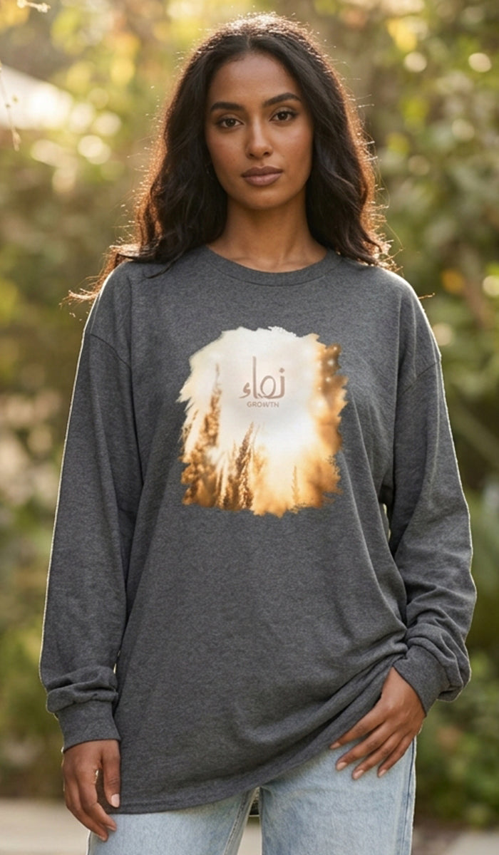 A woman wearing a dark heather gray long-sleeve t-shirt with a circular graphic of a golden sunset over a forest and the Arabic word 'Tawhid' in the center, paired with light blue jeans, set against a blurred outdoor background.