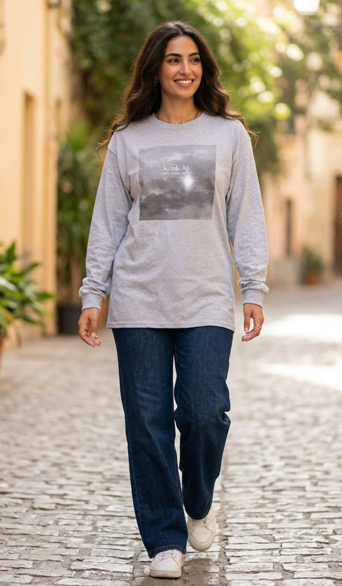 A woman walking down a cobblestone street wearing a light gray long-sleeve graphic t-shirt, dark blue jeans, and white sneakers.