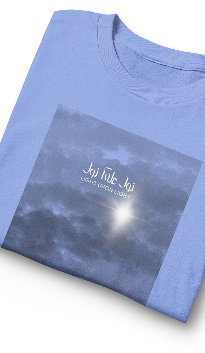 A folded light blue t-shirt featuring a square graphic print of a cloudy sky with a bright sunburst and the text 'LIGHT UPON LIGHT'.
