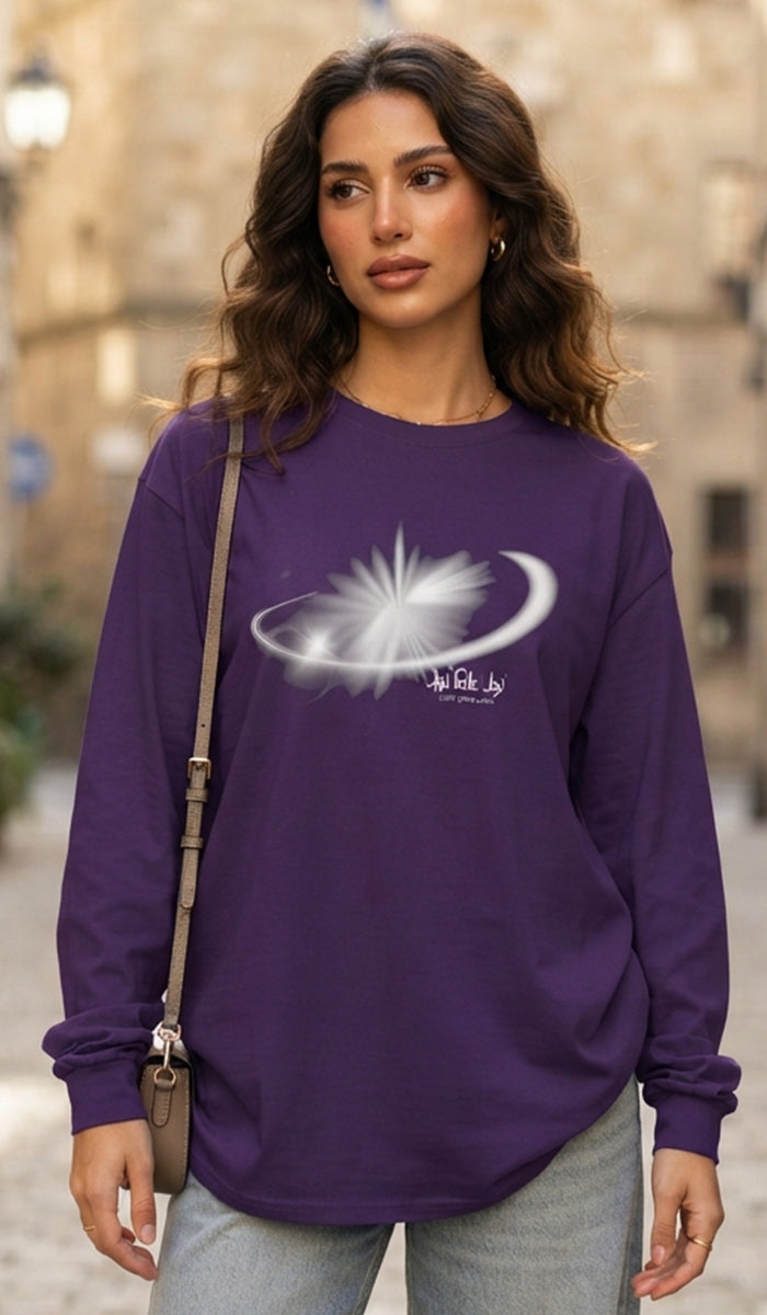 A woman wearing a purple long-sleeve t-shirt with a white crescent moon and star graphic on the chest, paired with light gray jeans and a brown shoulder bag, set against a blurred street background.