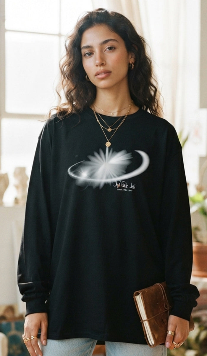 A woman wearing a black long-sleeve t-shirt with a white celestial graphic of a sun and crescent moon on the chest, holding a brown leather notebook in a softly lit indoor setting.