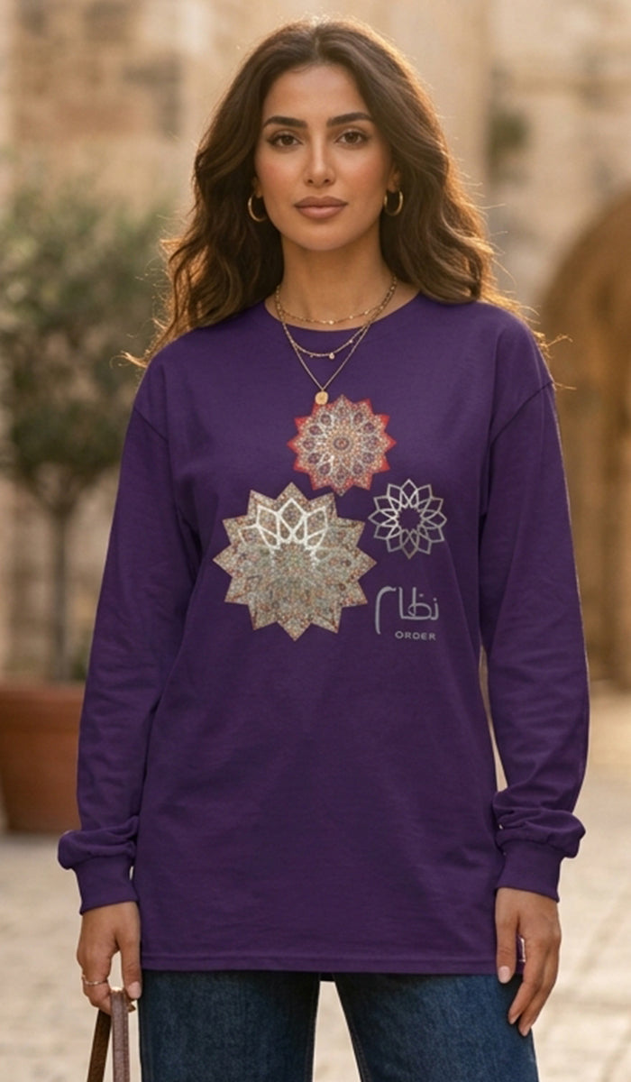 A woman wearing a purple long-sleeve t-shirt with three silver-toned mandala prints on the front, paired with blue jeans, set against a blurred outdoor background.