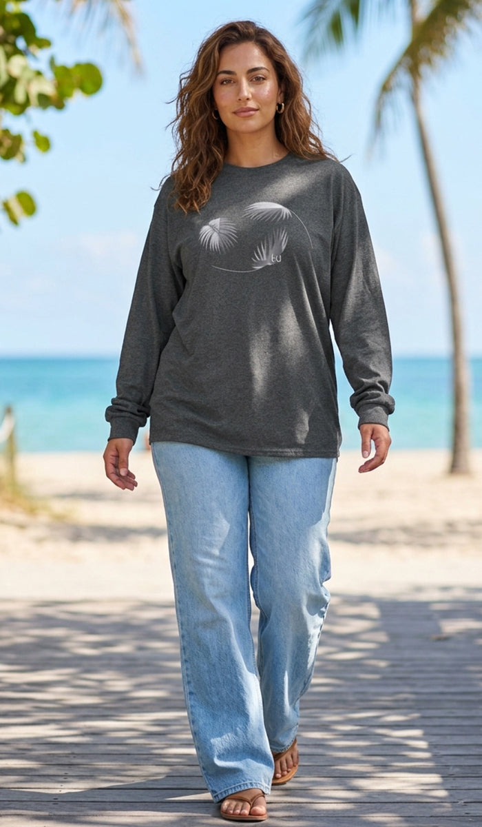 A woman walking on a wooden boardwalk at a beach, wearing a dark charcoal gray long-sleeve t-shirt with a white graphic print of two overlapping leaves, paired with light blue jeans.