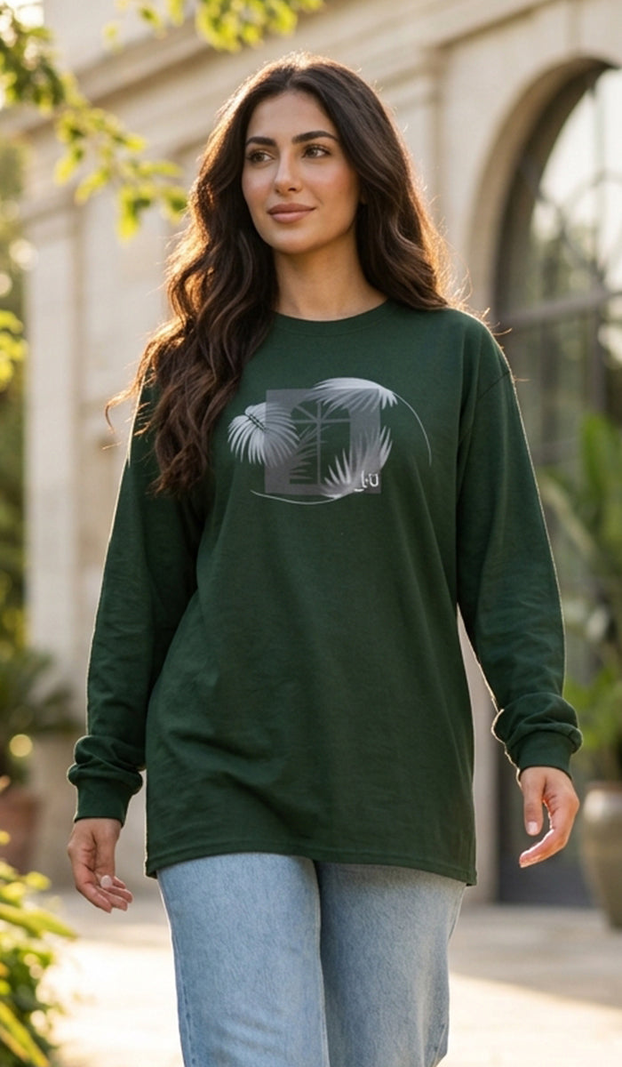 A woman wearing a dark green long-sleeve t-shirt with a gray graphic print of a house and palm leaves, paired with light blue jeans, standing outdoors in front of a stone building.