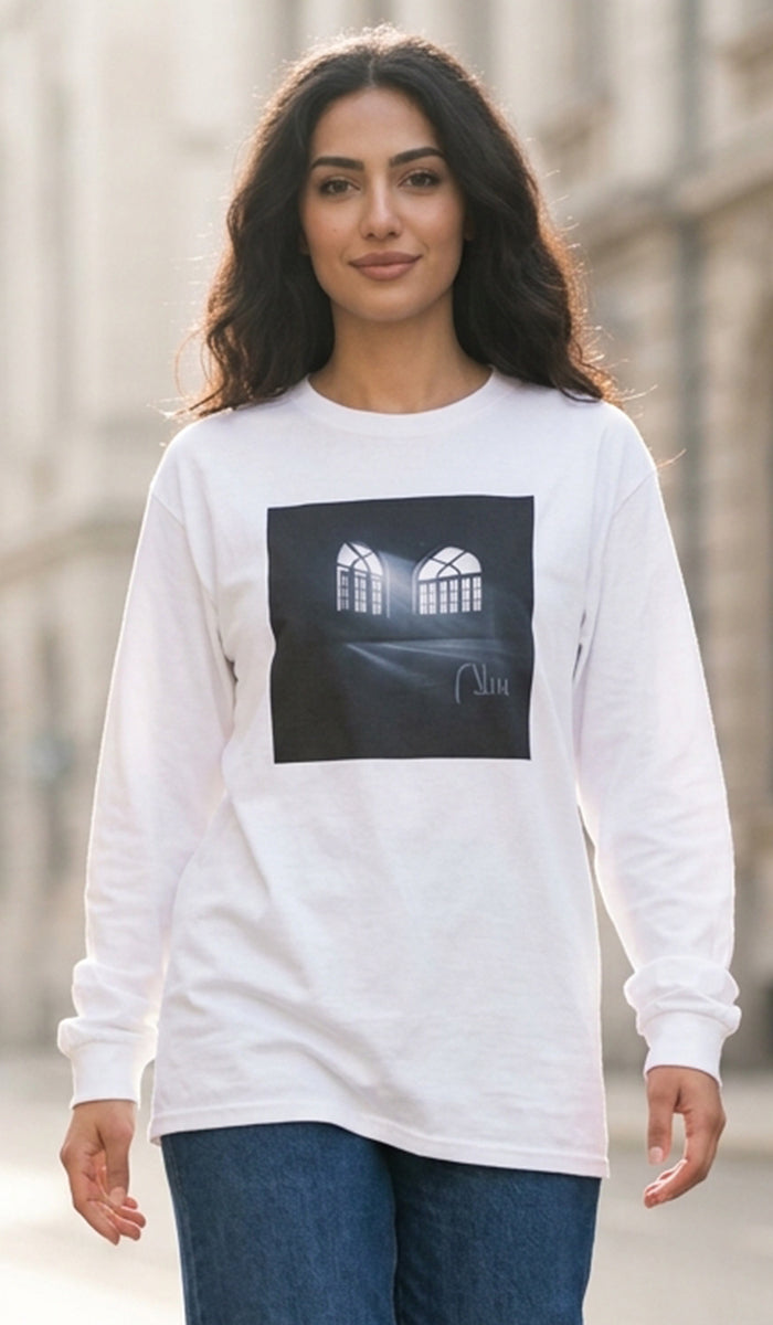 A woman wearing a white long-sleeve t-shirt with a black square graphic print of two arched windows, paired with blue jeans, standing outdoors against a blurred stone building background.