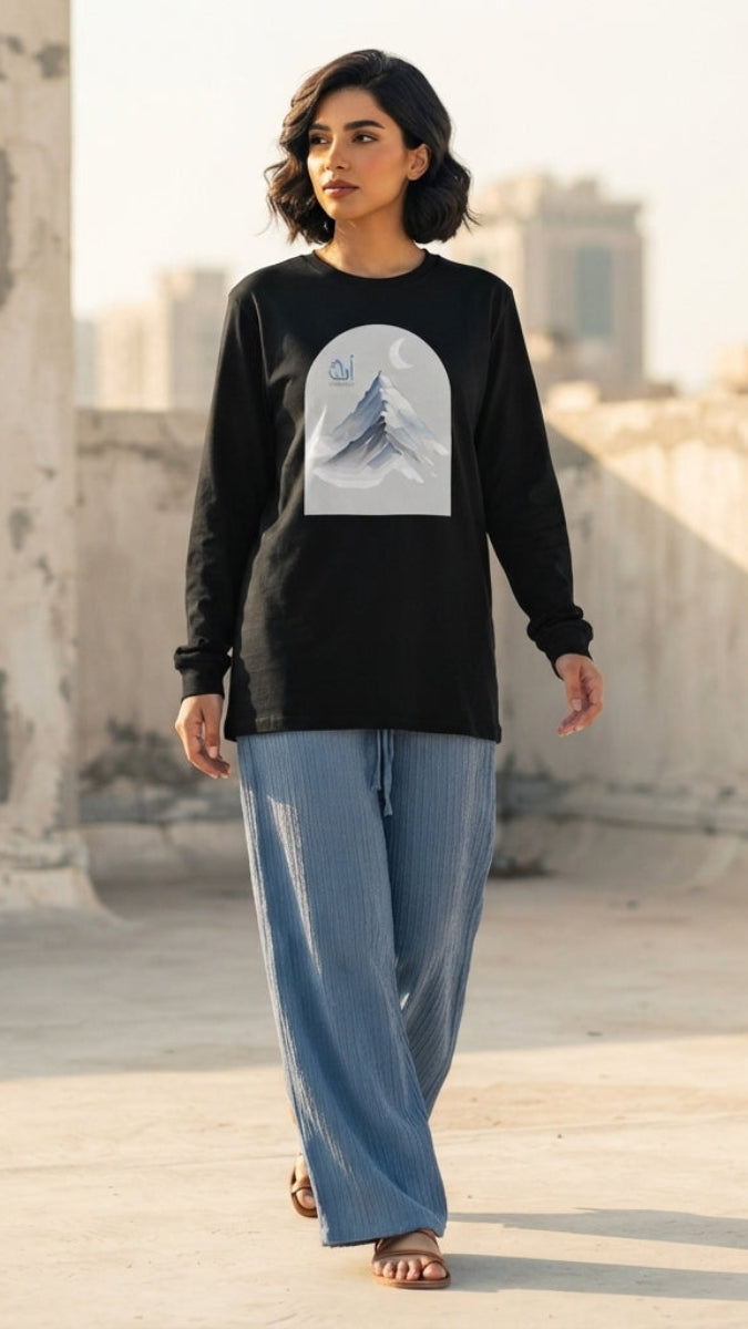 A woman stands on a sunlit rooftop wearing a black long-sleeve top with a large white arched graphic on the front and loose light blue pants.