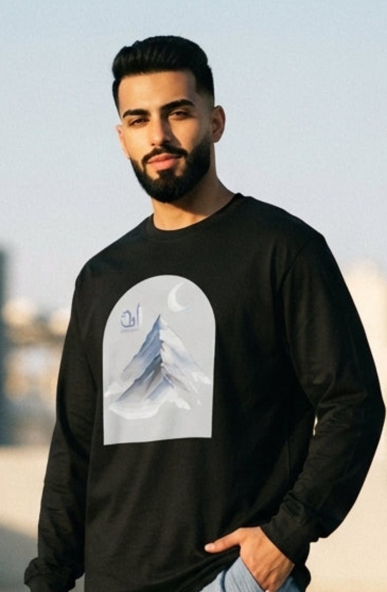 A man wearing a black long-sleeve crewneck sweatshirt with a white graphic print of a mountain under a crescent moon, paired with light blue wide-leg jeans and white sneakers, standing on a rooftop terrace.