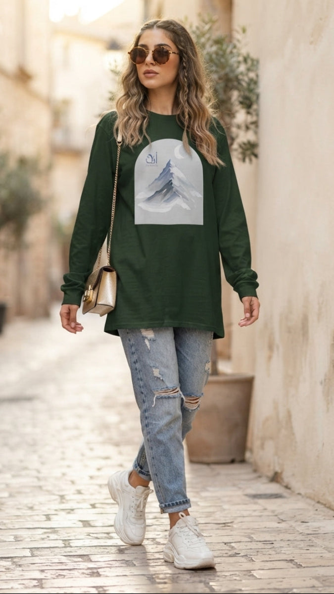 A woman walking down a sunlit cobblestone alley wearing a dark green long-sleeve tunic top with a large mountain graphic on the front, light-wash distressed jeans, white sneakers, and sunglasses.