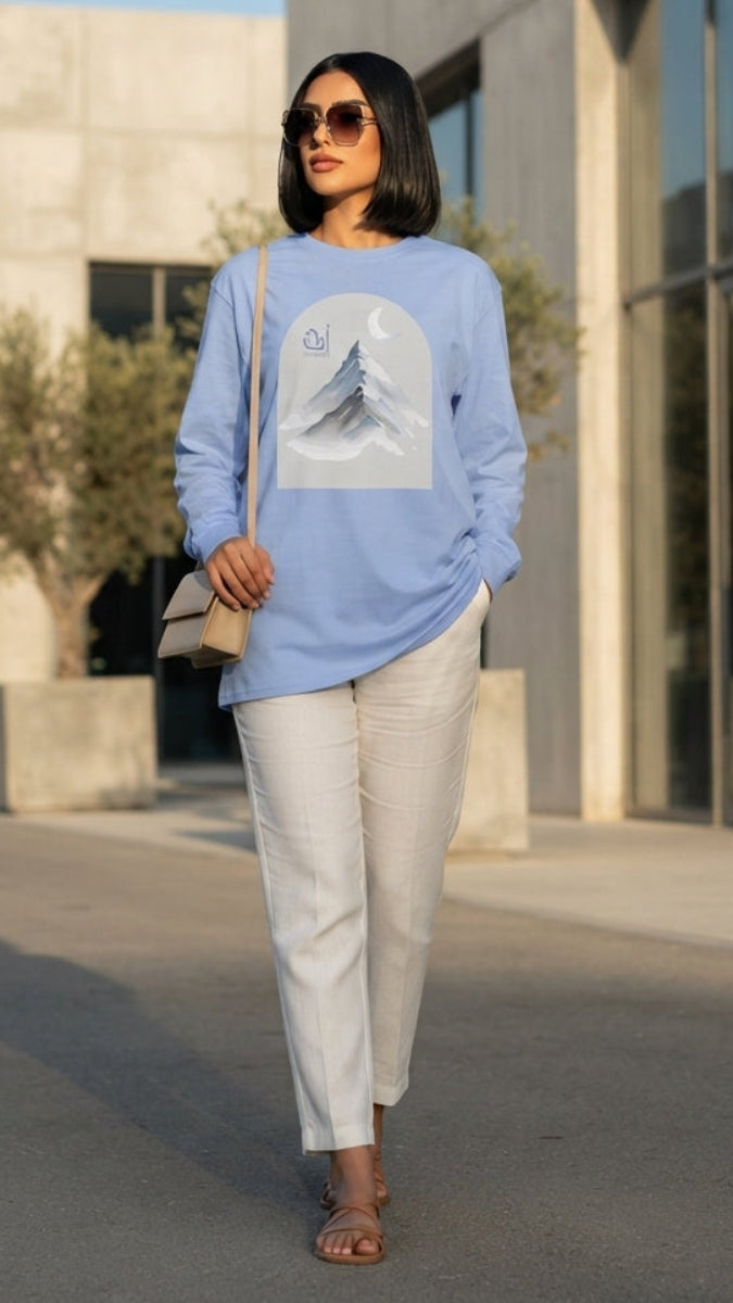 A woman walking outdoors in front of a modern concrete building, wearing a light blue long-sleeve sweatshirt with a large white graphic print on the front, paired with off-white pants and sandals.