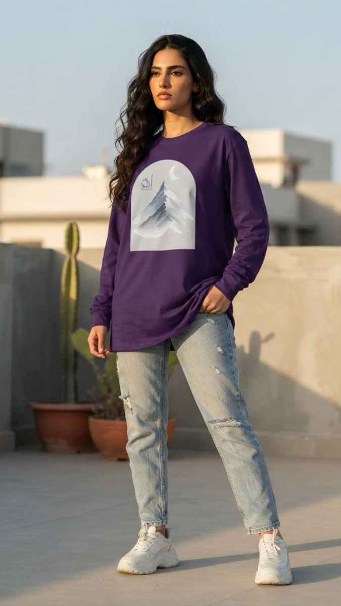 A woman stands on a rooftop wearing a purple long-sleeve t-shirt with a white graphic print, light-wash distressed jeans, and white sneakers.