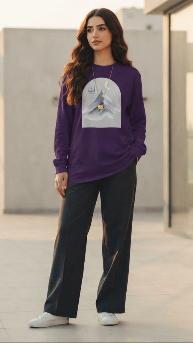A woman stands on a modern rooftop terrace wearing a purple long-sleeve sweatshirt with a light gray graphic print on the front, paired with black wide-leg pants and white sneakers.