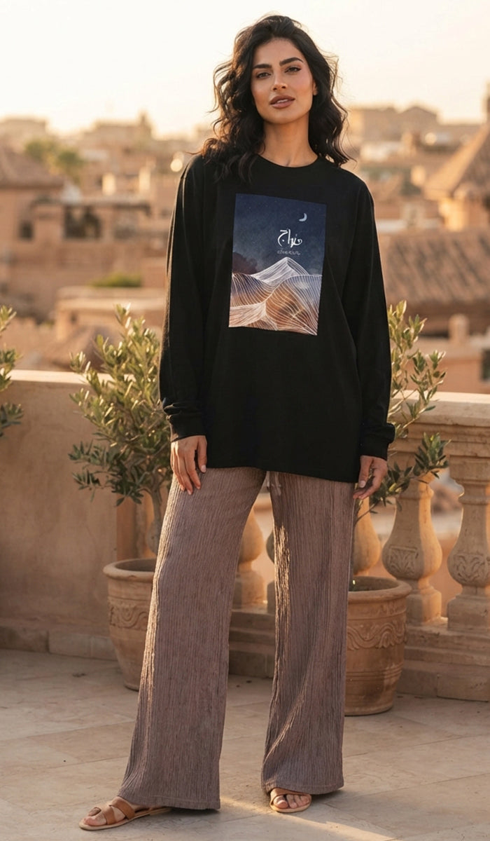 A woman stands on a sunlit terrace wearing a black long-sleeve top with a rectangular scenic graphic print on the front, paired with wide-leg taupe pants and sandals. The background shows warm-toned buildings and potted plants.