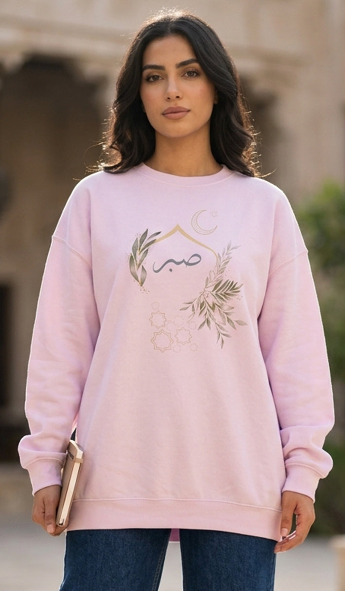 A woman wearing a light pink crewneck sweatshirt with a bohemian-style graphic print featuring a crescent moon, stars, and botanical elements, paired with blue jeans.