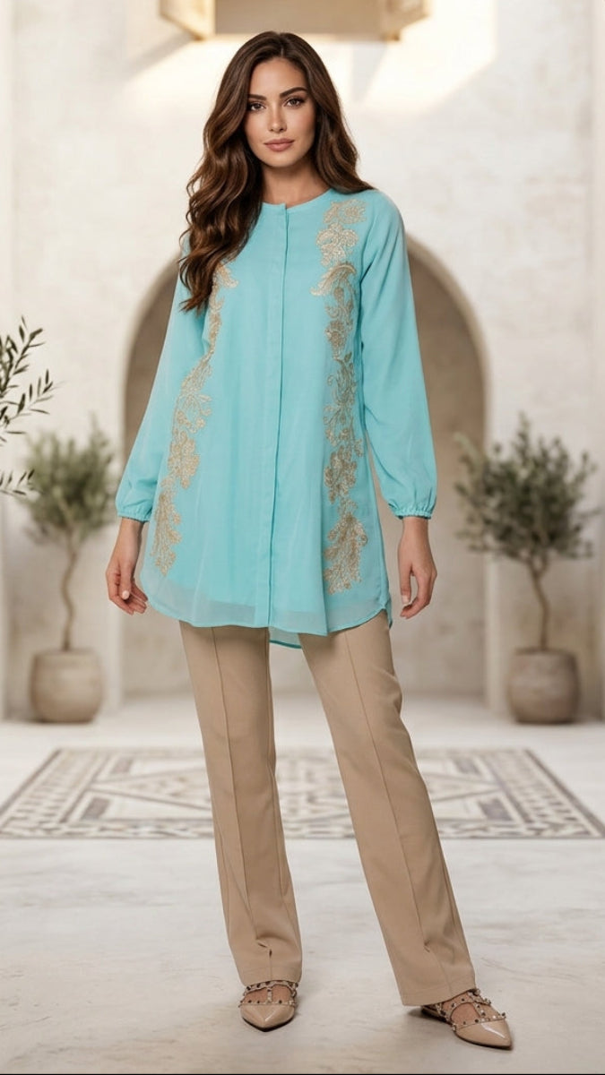 Woman wearing a light blue top with floral embroidery and beige pants in an indoor setting.