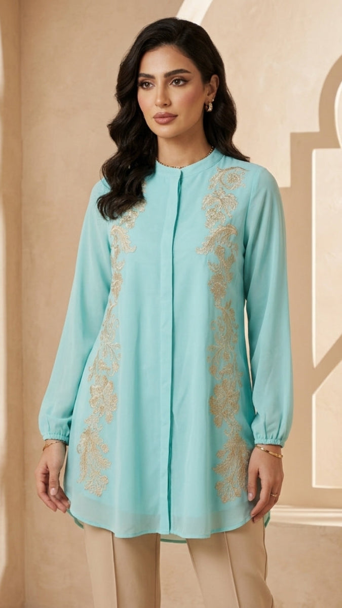 Woman wearing a light blue embroidered top with beige pants against a beige wall.