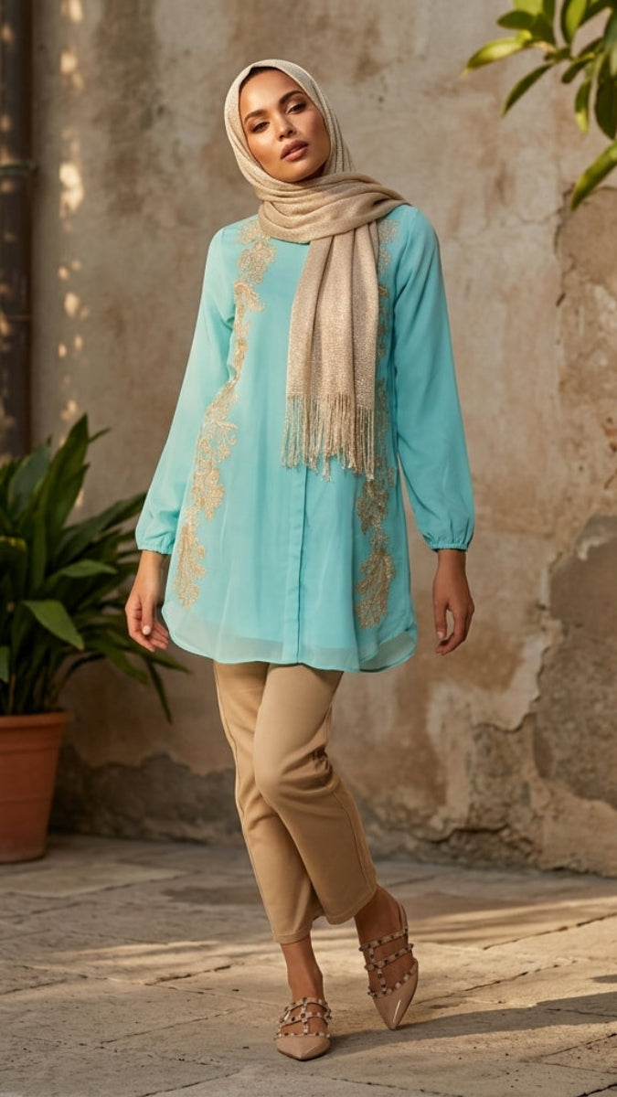 Woman in a light blue top and beige pants standing in front of a textured wall with plants.