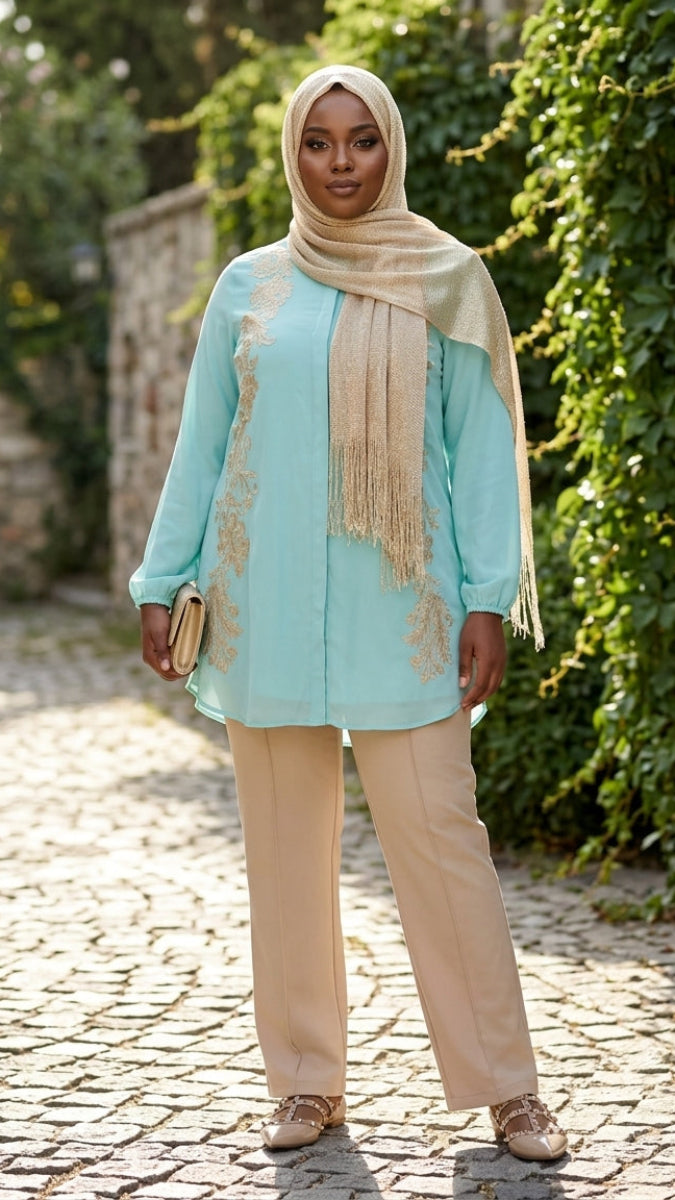 Woman in a light blue top and beige hijab standing on a cobblestone path with greenery.