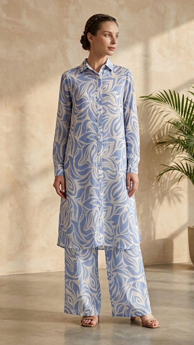 Woman wearing a blue patterned outfit standing in a room with a plant and neutral walls.