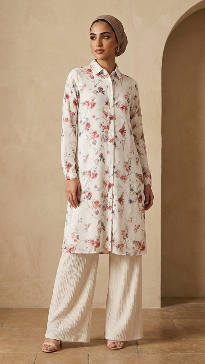 Woman wearing a floral long shirt and white pants against a beige wall.
