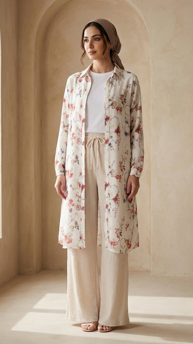 Woman wearing a floral long coat and beige pants in a sunlit room with arched walls.