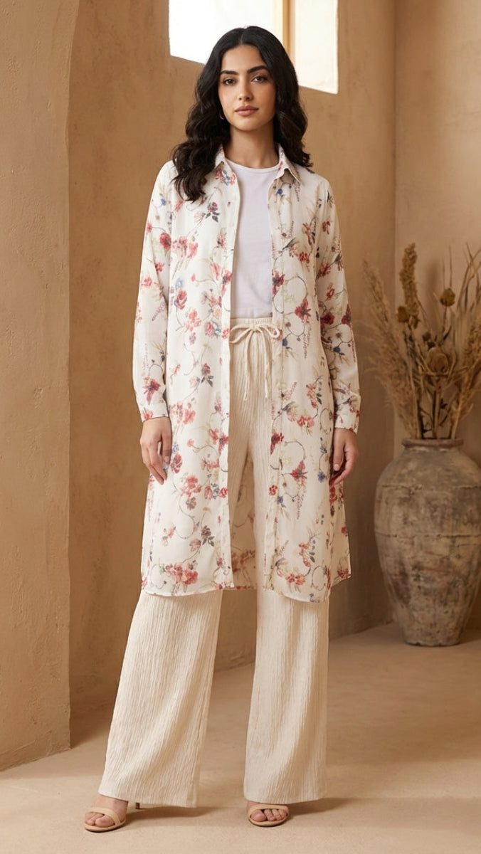 Woman wearing a floral long coat over a white shirt and pants in a warm-toned room.