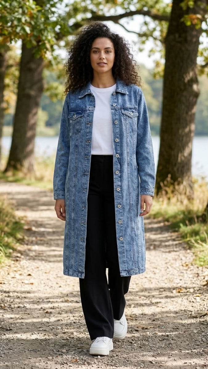 A woman walking on a tree-lined path outdoors, wearing a long light blue denim jacket over a white top and black pants.