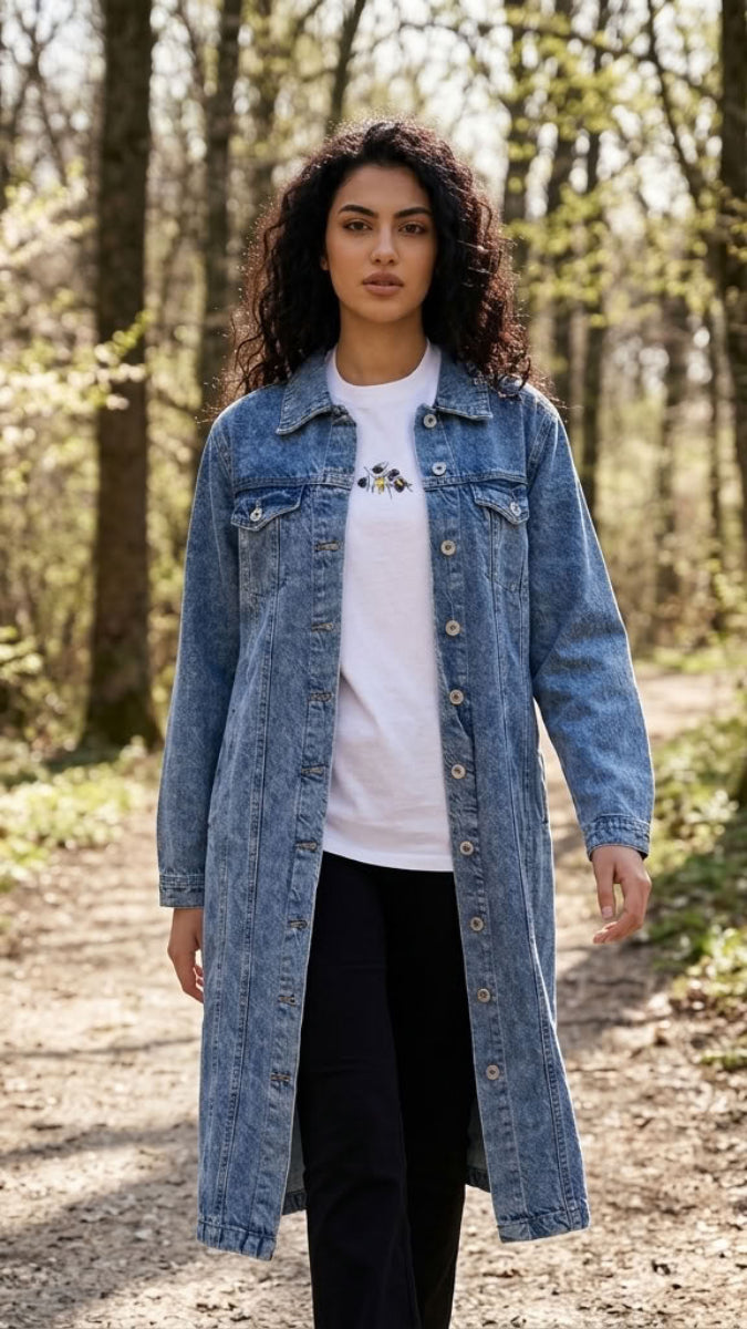 A woman stands on a wooded path wearing a long light blue denim jacket over a white graphic T-shirt and black pants.