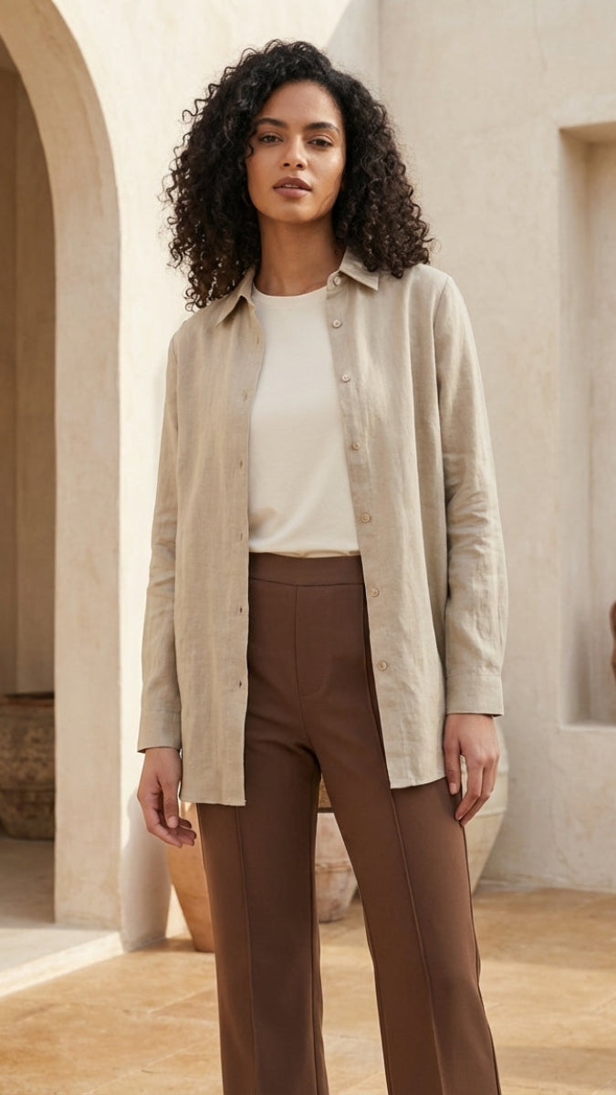 Woman wearing a beige jacket and brown pants standing in a sunlit room with architectural elements.